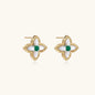 Gold quatrefoil stud earrings with white mother-of-pearl petals and emerald green gemstone center on beige background.